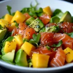 Salade de saumon fumé avec morceaux de mangue, avocat, jeunes pousses et graines de sésame dans une assiette blanche.