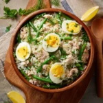 Salade de riz, thon, haricots verts et œufs servie dans un grand saladier, accompagnée d'herbes fraîches.