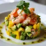 Tartare de crevettes, avocat et mangue servi dans un cercle, garni de coriandre fraîche, sur une assiette blanche.
